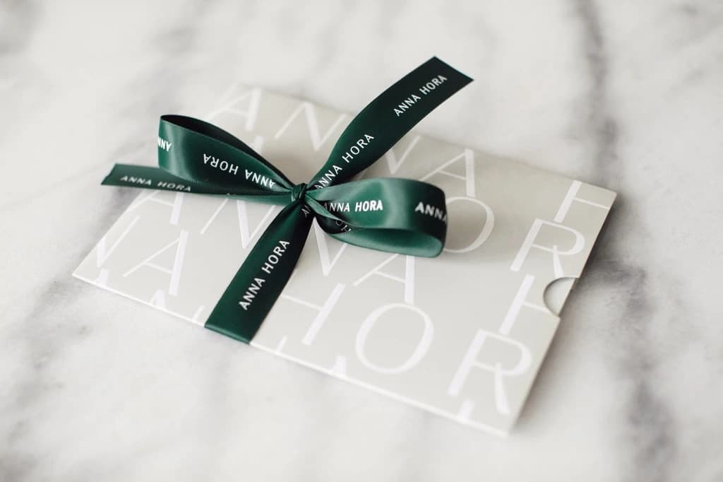 An ANNA HORA gift card tied with a branded green satin ribbon on marble table top