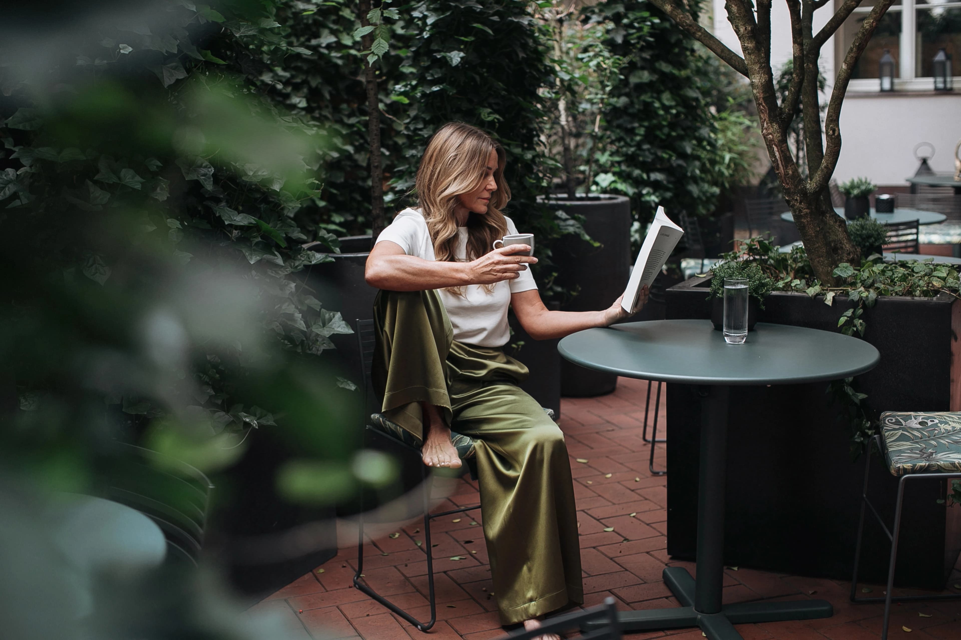 A woman reads a book outdoors in green silk trousers, coffee in hand.