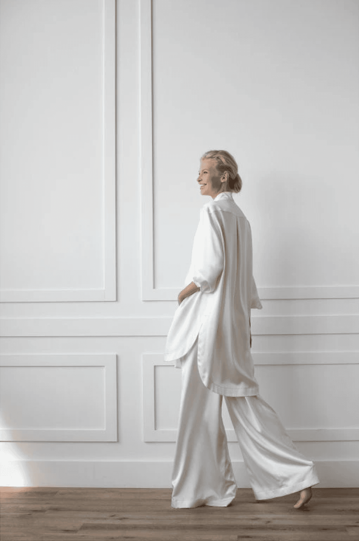 A woman walks in an off-white silk lounge set against a white panelled wall.