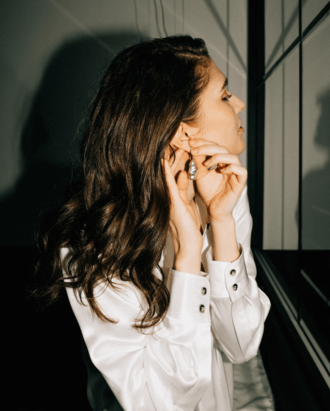 A woman in a white silk lounge shirt puts on an earring, side profile.
