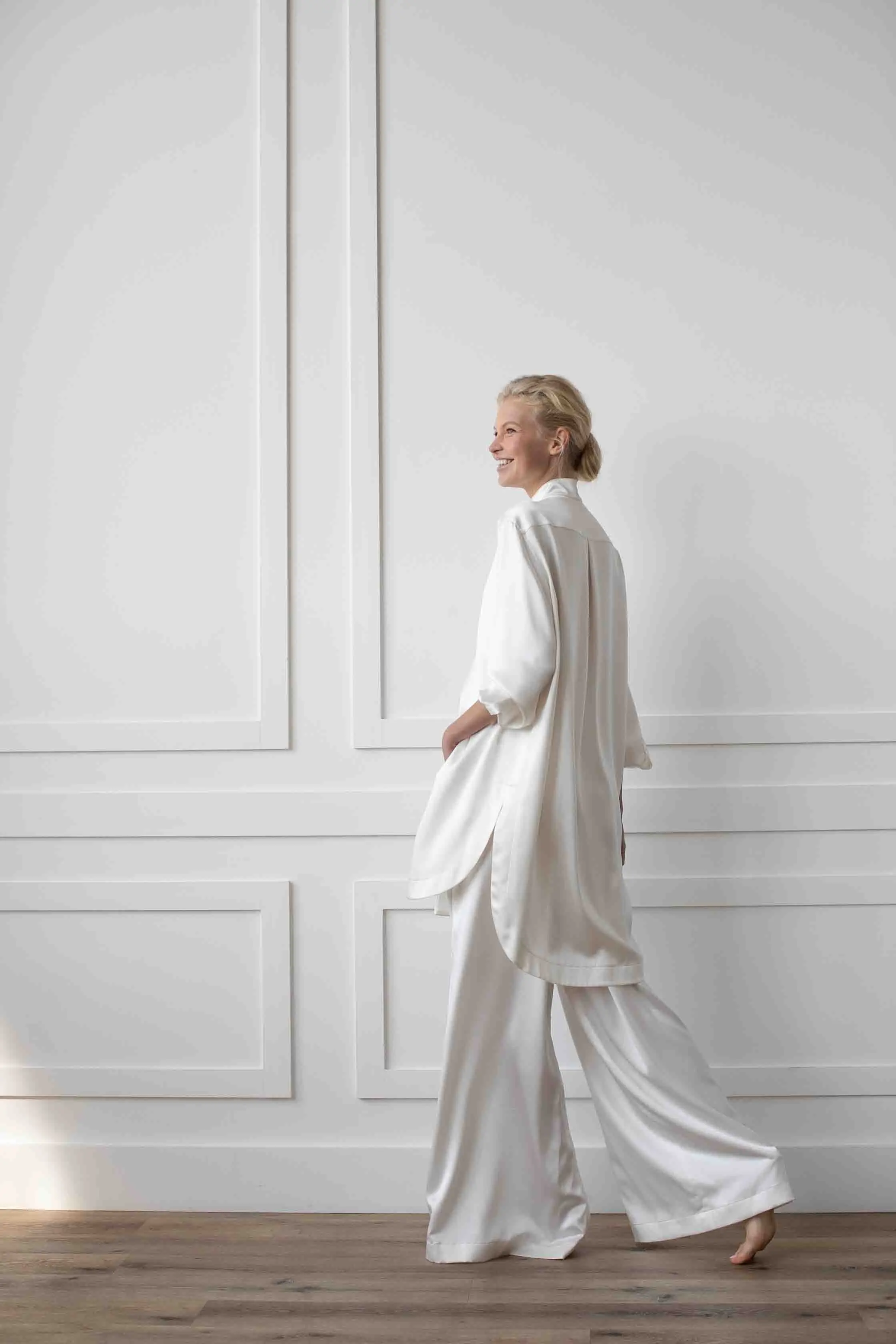 A woman walks in an off-white silk lounge set against a white panelled wall.
