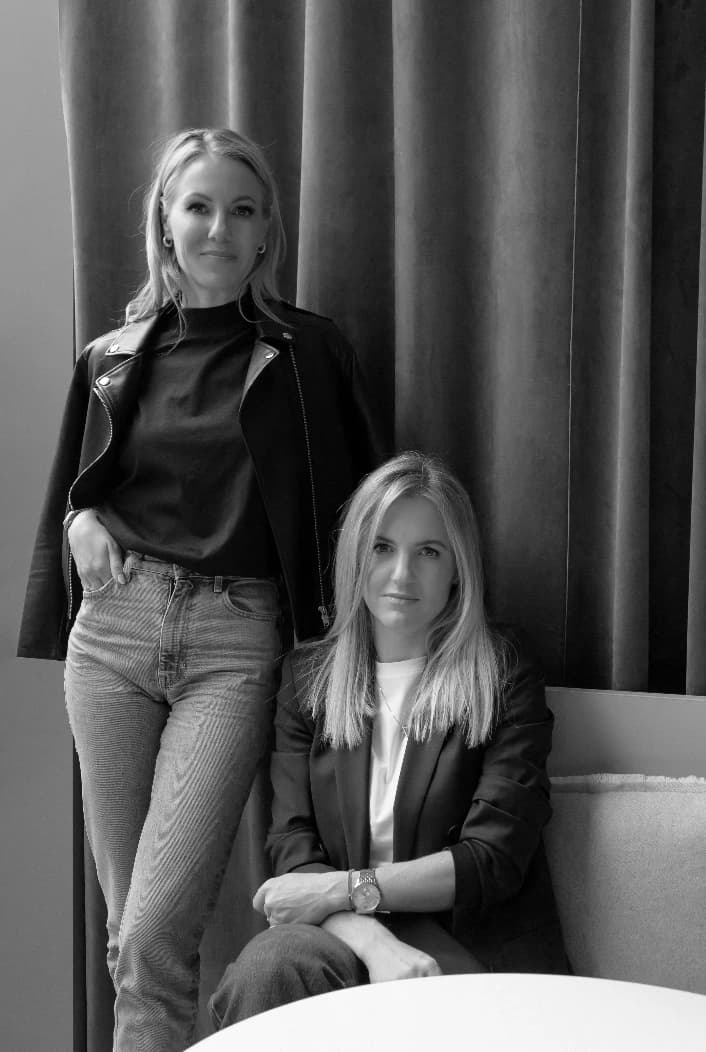 Two women, one standing and one seated, photographed in black and white against draped curtains — the founders of ANNA HORA, Kat and Barbora.